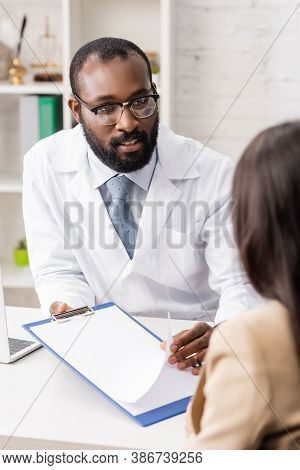 Selective Focus Of African American Doctor Talking To Patient While Holding Clipboard With Blank Pap
