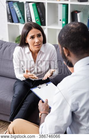 Selective Focus Of Tense Woman Gesturing While Talking To African American Psychologist Holding Clip