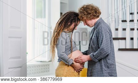 Young Hipster Pregnant Couple Standing Opposite Each Other In Contemporary Apartment And Tenderly St
