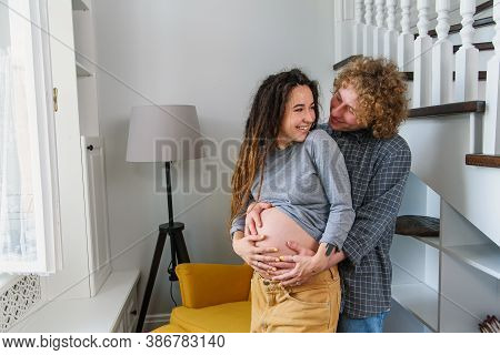 Happy Hipster Pregnant Couple Standing Together Near Stairs In Contemporary Apartment And Tenderly T
