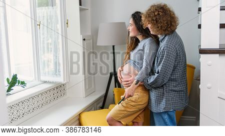 Happy Hipster Pregnant Couple Standing Together Near Window In Contemporary Apartment And Tenderly T