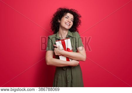Cute Woman In A Dress And Curly Hair Embracing A Present And Smile On A Red Wall At Studio