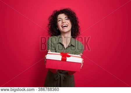 Charming Brunette Woman With Curly Hair Giving A Present At Camera Smiling On A Red Studio Wall