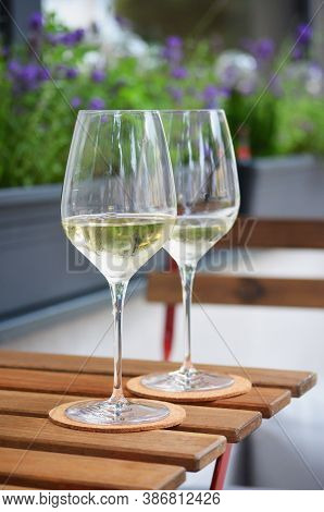 Close Up Of Two Glasses Of White Wine Standing On A Wooden Table, Flower Pot In Background