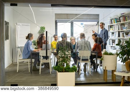 Confident mature businessman giving a presentation to his crative team in office. Happy leadership man training young businessmen and smiling businesswomen sitting at conference table.  Business brief