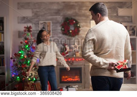 Young Man Holding A Gift Behind Him For His Girlfriend On Christmas Day Wearing Matching Clothes.