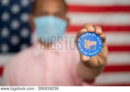 Man In Medical Mask Showing I Voted By Mail Sticker With Us Flag As Background - Concept Of Mail In 