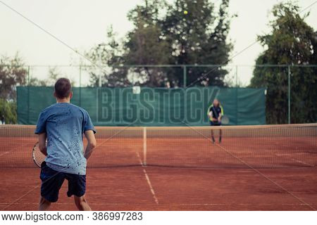 Man In Blue Shirt And Blue Pants Seen From Behind Playing Tennis, Wide Open Clay Orange Playing Fiel