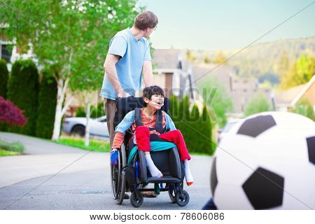 Caucasian Father Helping Disabled Biracial Son In Wheelchair Play Soccer Outdoors On Street