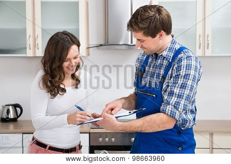 Woman Writing On Clipboard With Plumber In Kitchen Room