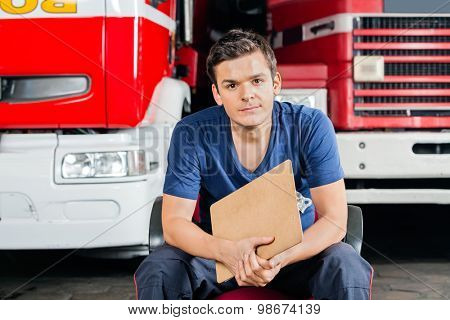 Portrait of confident male firefighter holding clipboard while sitting against firetrucks at station