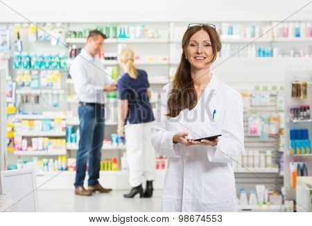 Portrait of smiling female chemist holding digital tablet while assistant and customer standing in background at pharmacy
