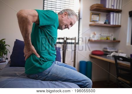 Side view of male patient grimacing from back ache while sitting on bed at hospital ward