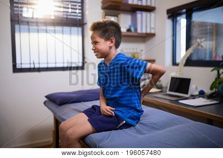 Boy frowning with backache while sitting on bed at hospital ward