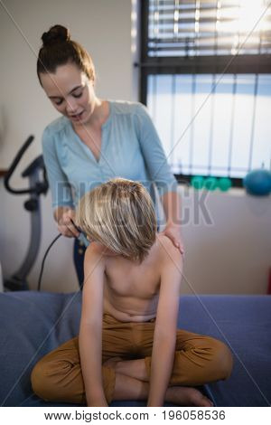 Female therapist scanning shoulder of shirtless boy with ultrasound machine at hospital ward