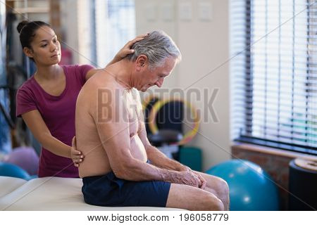 Young female therapist giving back massage to senior male patient sitting on bed at hospital ward