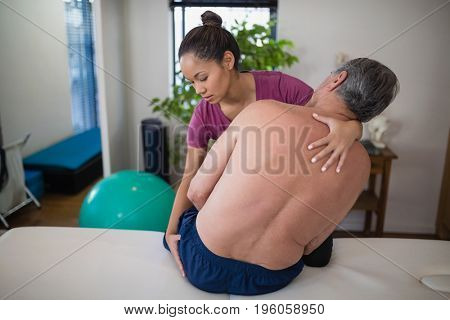 Young female therapist examining buttocks of shirtless senior male patient sitting on bed at hospital ward