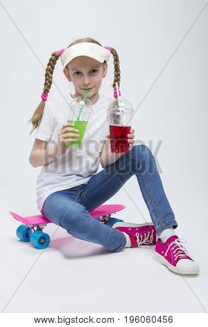 Kids Concepts. Portrait of Little Caucasian Blond Girl in Visor Sitting on Pink Pennyboard With Two Cups of Juice and Drinking Through Straw. Against White. Vertical Image