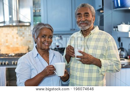 Portrait of smiling senior couple with coffee cups standing in kitchen at home