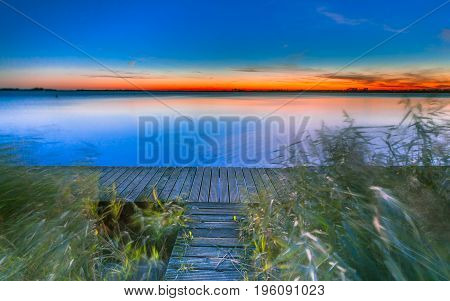 Long Exposure Image Of Blue And Orange Sunset Over A Pier On The Shore Of A Lake