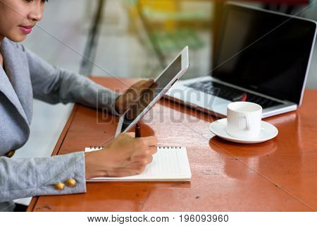 Beautiful Asian businesswoman was looking at the tablet and taking notes on her desk on the wood table in her office. Finance Banking and Technology Concepts.
