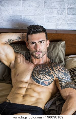 Shirtless sexy muscular male model lying alone on his bed in his bedroom, looking at camera with a seductive attitude