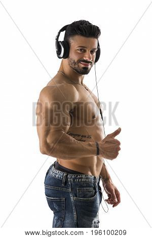 Handsome shirtless muscular bodybuilder man listening to music with headphones, isolated on white background, while doing thumb up sign with one hand