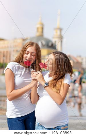 Lesbians mothers, pregnant couple, happy samesex family in the city park at summer. Women eating sweets, heart shaped candies, symbol of love