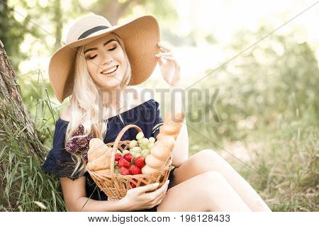 Smiling blonde girl 22-24 year old holding basket ful of fresh food i park. Having picnic in park. Summer season.