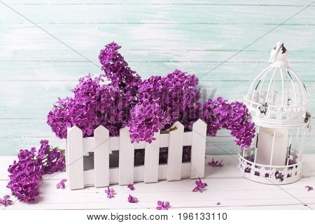 Shabby chic flowers composition. Fresh lilac flowers in box and l candleholder with candle in form of cage on white wooden background against turquoise wall. Selective focus.
