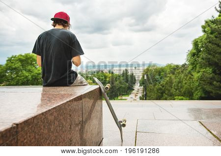 The long-haired skater in a cap and T-shirt sits with his back to the camera and thinks next to the skateboard against the background of the landscape landscape.