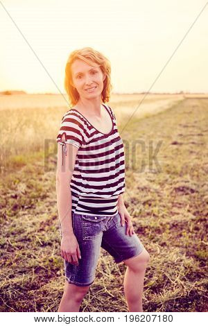 Portrait of young caucasian woman standing at summer sunny field at sunset. She is wearing casual clothes looking into camera smiling.