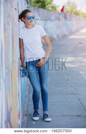 Hipster girl wearing blank white t-shirt, jeans and sunglasses posing against rough street wall, minimalist urban clothing style, mockup for tshirt print store