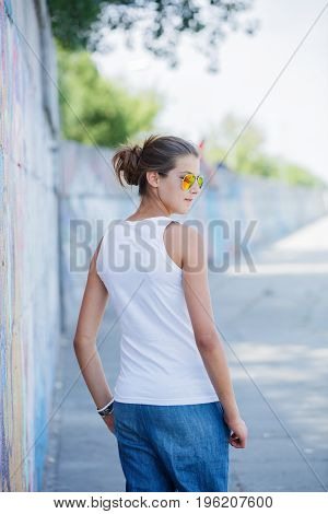 Hipster girl wearing blank white t-shirt, jeans and sunglasses posing against rough street wall, minimalist urban clothing style, mockup for tshirt print store