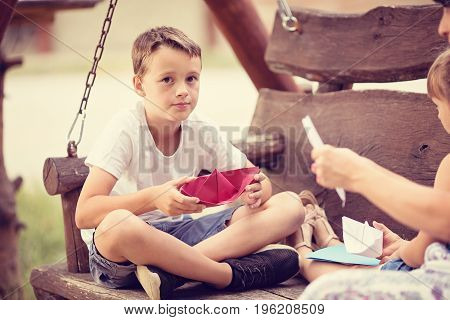 Three member caucasian young family sitting on a wooden swing outdoors on summer day folding a paper boats together.Focus is on boy who is holding a red paper boat and looking into camera.