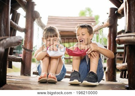 Two young caucasian children sitting next to each other with bent knees in a wooden house outdoors on sunny summer day playing with paper boats