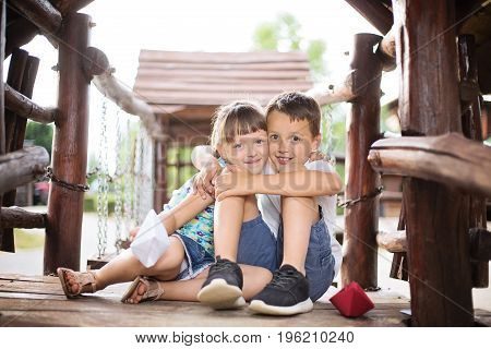 Two young caucasian children sitting next to each other in a wooden house hugging. Photo is taken outdoors on sunny summer day