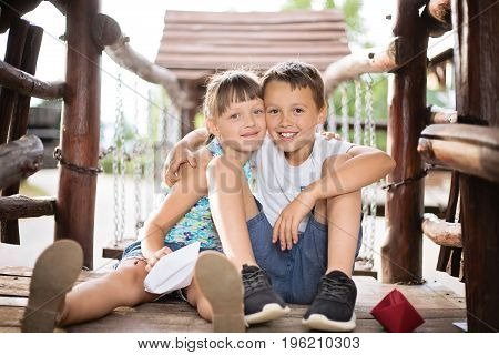 Two young caucasian children sitting next to each other in a wooden house hugging. Photo is taken outdoors on sunny summer day