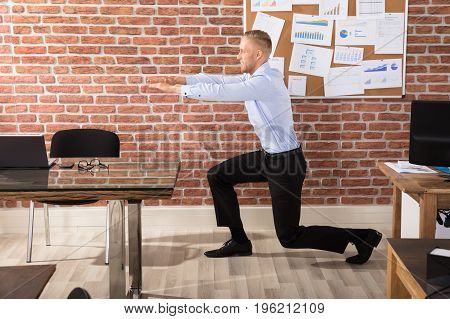 Attractive Businessman Doing Exercise In Office Against Brick Wall