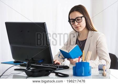 Young Confident Businesswoman Looking At Blue Diary Using Computer At Desk In Office