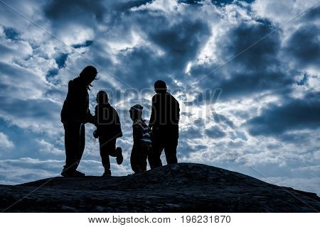 Silhouette back view of happy and friendly family. Father and mother with children hikers enjoying view at the top of a mountain against blue sky with dark cloudy in a winter's day. Travel on vacation