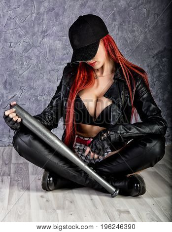 Stylish hipster woman with big tits in baseball cap and black jacket sitting on a floor, holding baseball bat