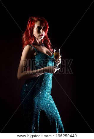 Young redhead sexy woman in blue elegant dress holding wineglass with champagne and poses in a dark