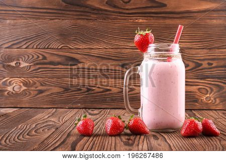 A tasty strawberry smoothie in a transparent mason jar on a dark wooden background. A single vegetarian pink milkshake with a red strawberry on the top and freshly grown red berries.