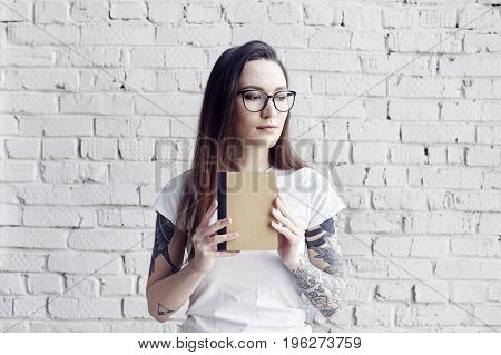 Pretty hipster tattooed woman poses in white t-shirt with book in hands isolated on white brick wall Hold a book in hand space for design layout