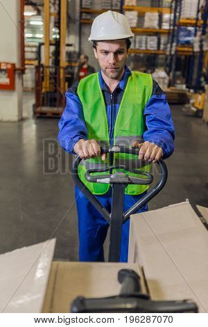 Man with stubble wearing uniform and hardhat steering pallet truck carrying boxes in warehouse.