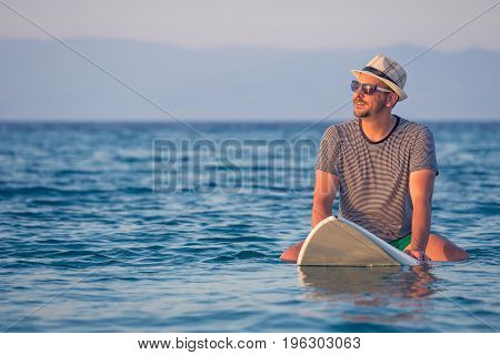 Man in hat and sunglasses sitting on surfboard in ocean. Summer vacation and travel concepts.