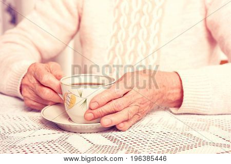 An elderly woman drinks tea at home. Senior woman holding cup of tea in their hands at table close-up