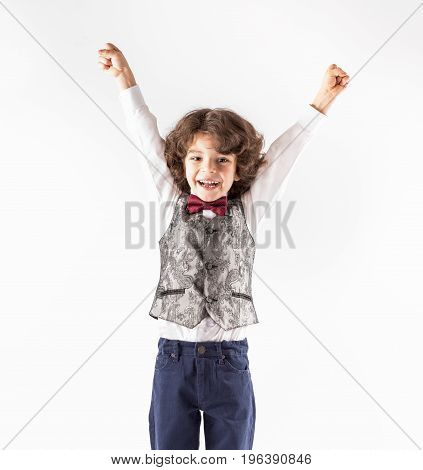 Cute cheerful curly-haired boy in a waistcoat raised his hands with joy. Close-up. Gray background.