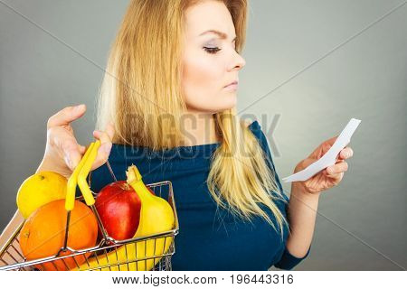 Worried woman holding shopping basket with fruits looking at bill receipt being scared of huge prices